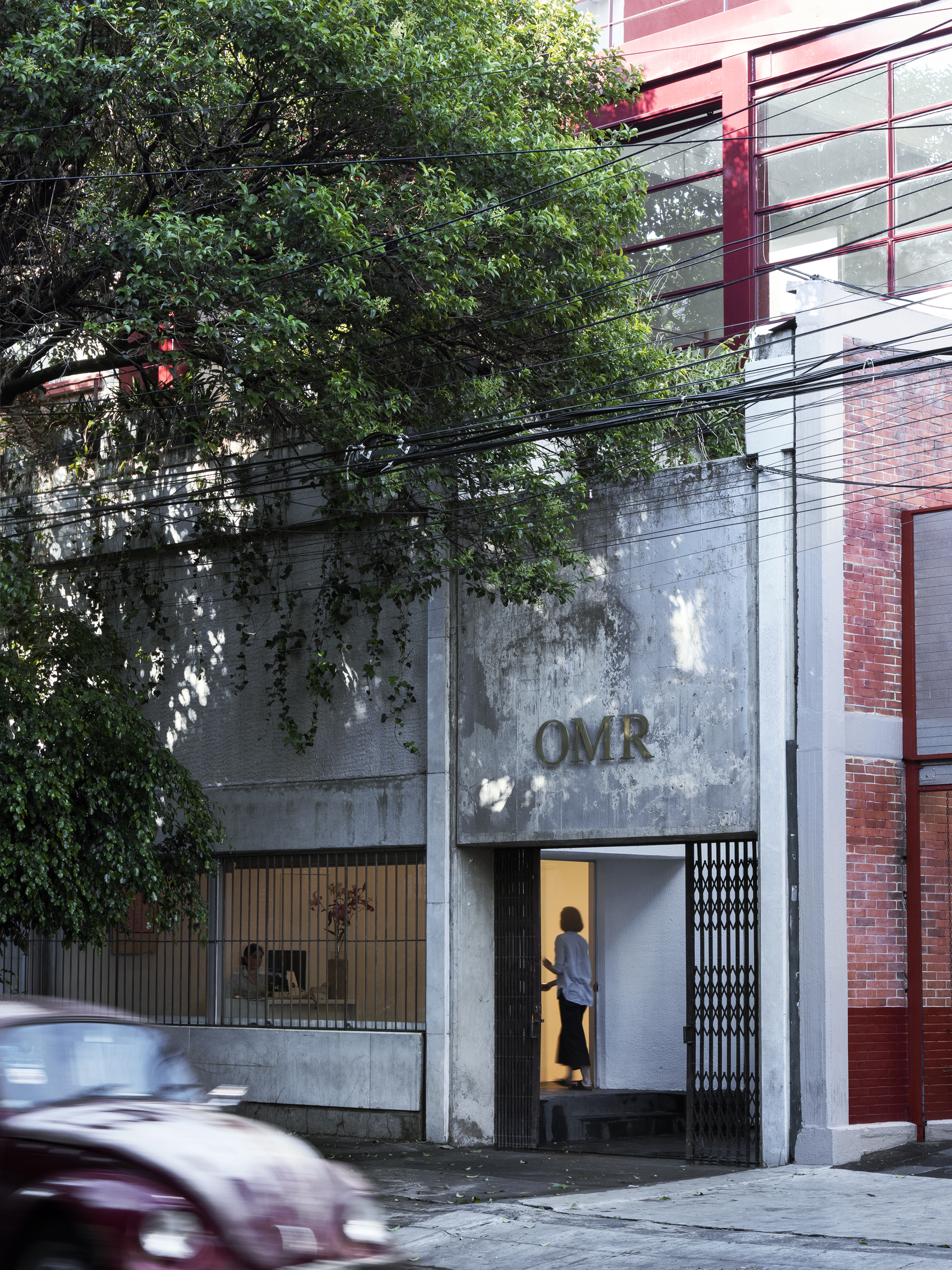 The bare cement and red iron facade of a gallery space in Mexico City lets the warm glow inside of its room out via a minimalist door overlooking a tree-filled street onto which a vintage car in the same shade of red is passing.