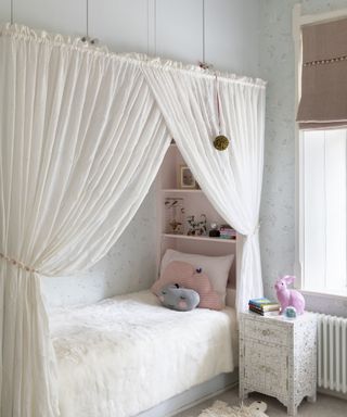 A child's bedroom with pale blue floral wallpaper and a recessed bed framed by white curtains