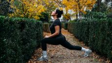 A woman performs a lunge outside. She wears leggings, a t-shirt and sneakers. Her left leg is in front and bent at a right angle, while her right angle is straight behind her. There are green leafy hedges either side of her and behind her we see trees with yellow leaves.