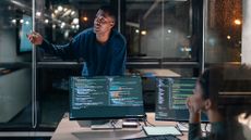 Male and female software developers discussing team strategy in an open plan office space with computer code pictured on desktop monitors.