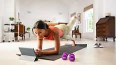 woman facing ipad device doing pilates move from plank position in living room setting wearing pink vest and light leggings