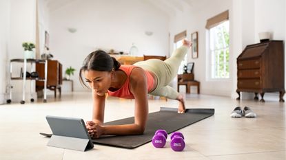 Woman facing tablet doing Pilates move from plank position in living room setting wearing pink vest and light leggings