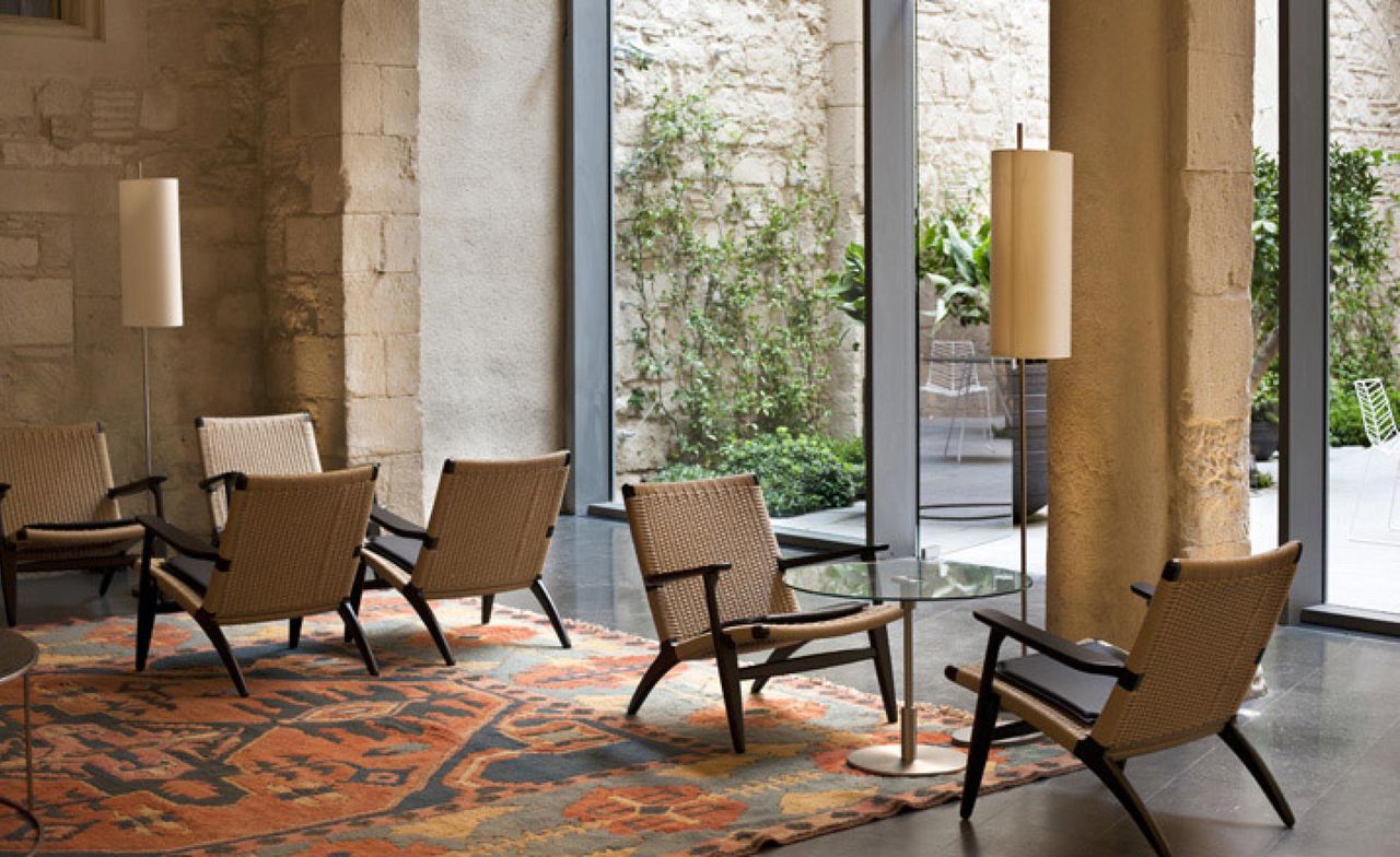 A hotel sitting area with wood and wicker chairs, round glass coffee tables, floor lamps, a patterned rug, rugged walls and large glass windows.