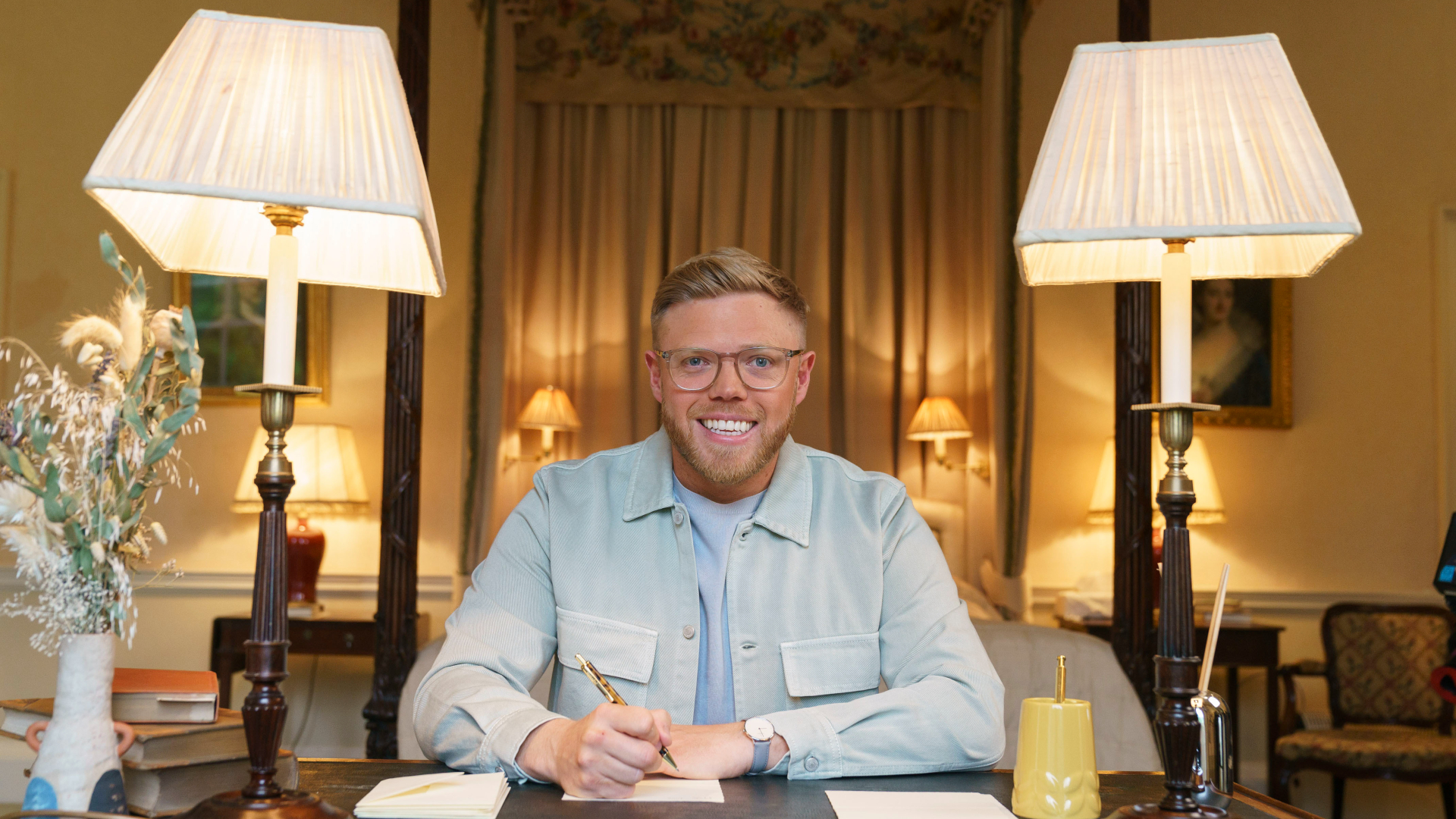 Rob Beckett sits at a desk bordered by two lamps.