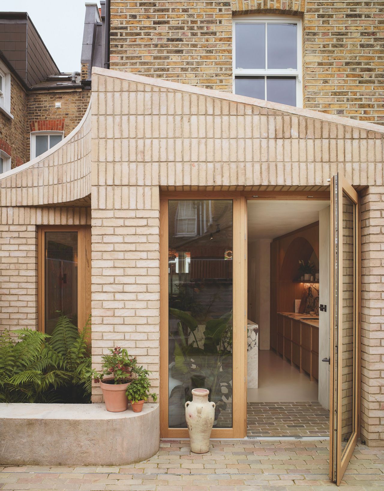 An exterior doorway leading to a kitchen extension with a curved pointed brick surround and a large pot sits next to the glass next to the wood-framed door