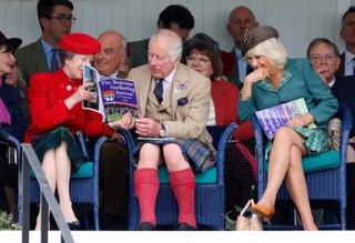 Princess Anne wearing a red coat sitting in a chair next to King Charles and Queen Camilla reading a program at the Braemar Gathering Highland Games in Scotland