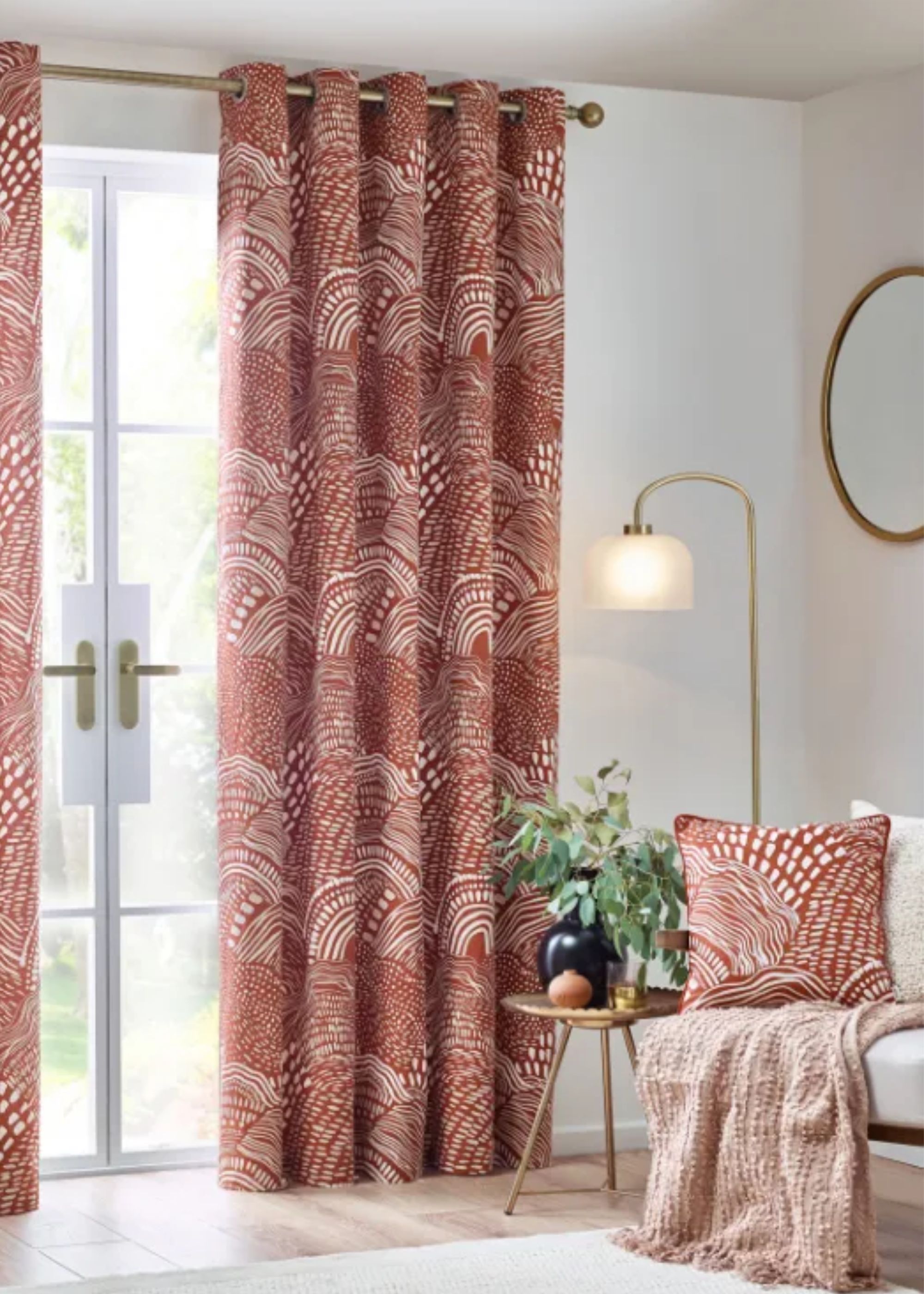 Red colored curtains in a living room and beside it is a side table with a plant and vase. There is also a section of the sofa in view with a cushion and throw on top