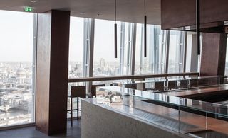 Oblix, London, UK. A bar area with a square glass topped counter, long pendant lights and a long table with high chairs against a window with a view the the city below.