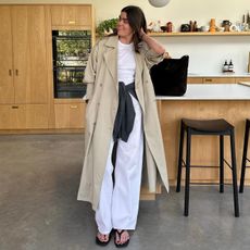 British style influencer Anna Newton of The Anna Edit poses in her stylish kitchen wearing a spring 2026 outfit idea with a trench coat, white T-shirt, a grey sweater around the waist, white cotton relaxed pants, and black flip-flop sandals