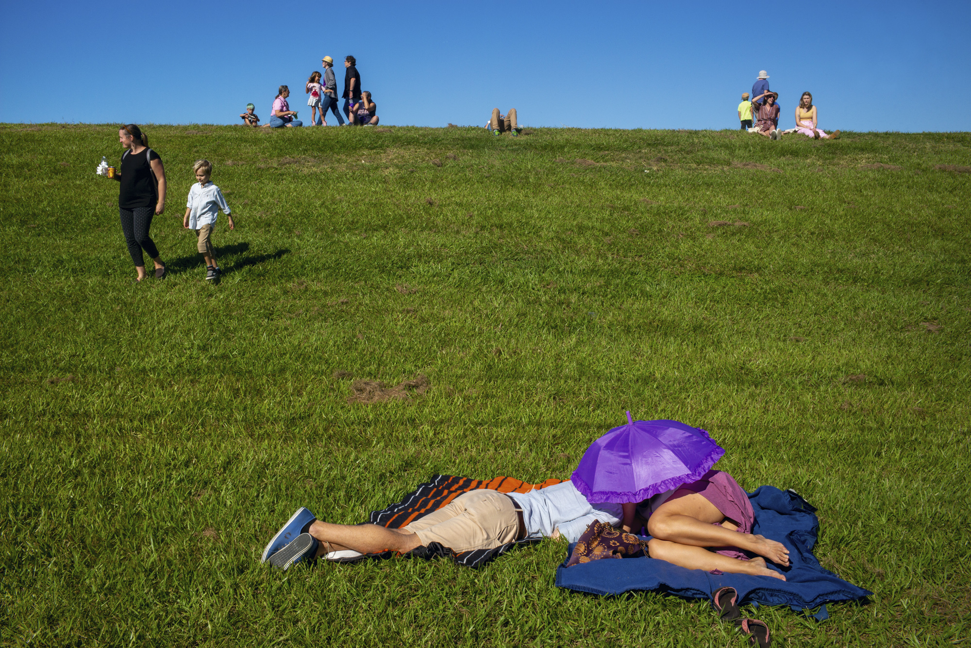 Two people lie asleep on blankets on a steep grassy bank under a frilly purple parasol, while a woman and boy walk down the slope