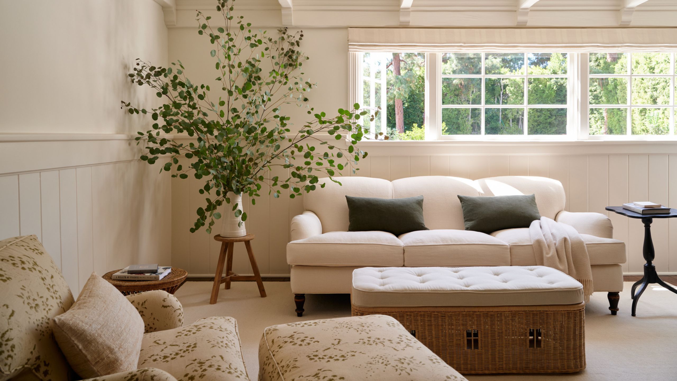 a warm neutral primary bedroom with half-height wainscoting and a sweet sitting area with white couch, ottoman and floral armchair