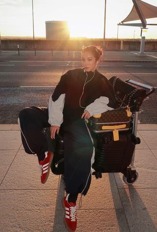 A winter travel outfit is shown in a photo of a woman sitting on top of her luggage at an airport terminal wearing reading glasses, headphones, a matching black tracksuit, and red suede sneakers