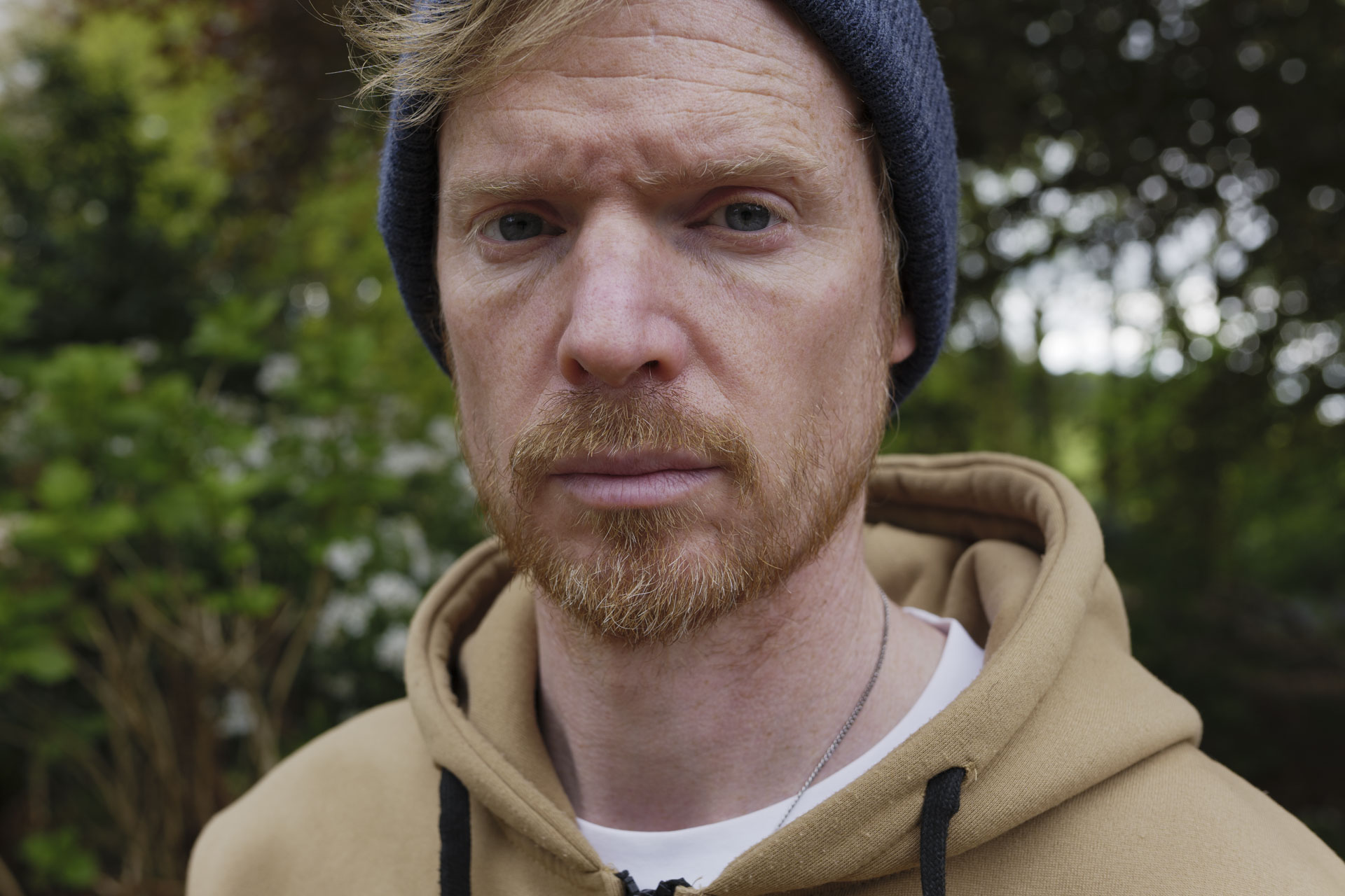 Portrait of a man in a beanie hat and hoodie, the background is lush green bushes and trees with dappled light