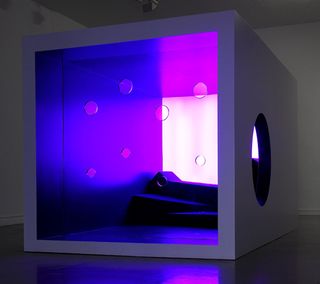 Purple Box, by Anna + Peter, 2010. A grey cube with a circular hole on the side, a glass front with round holes in it and purple lights glowing around it.