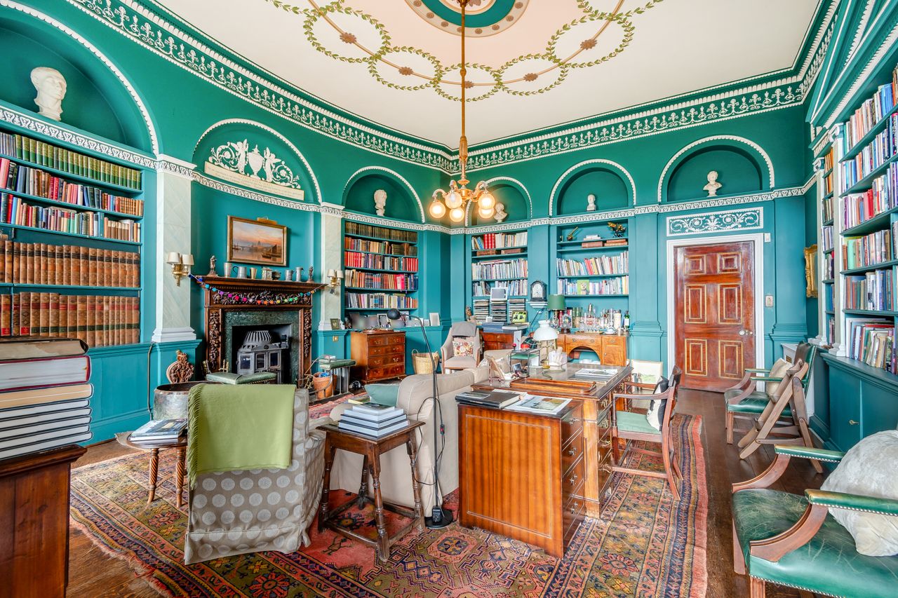 A teal library with an ornate ceiling and many colourful books and pieces of furniture scattered about