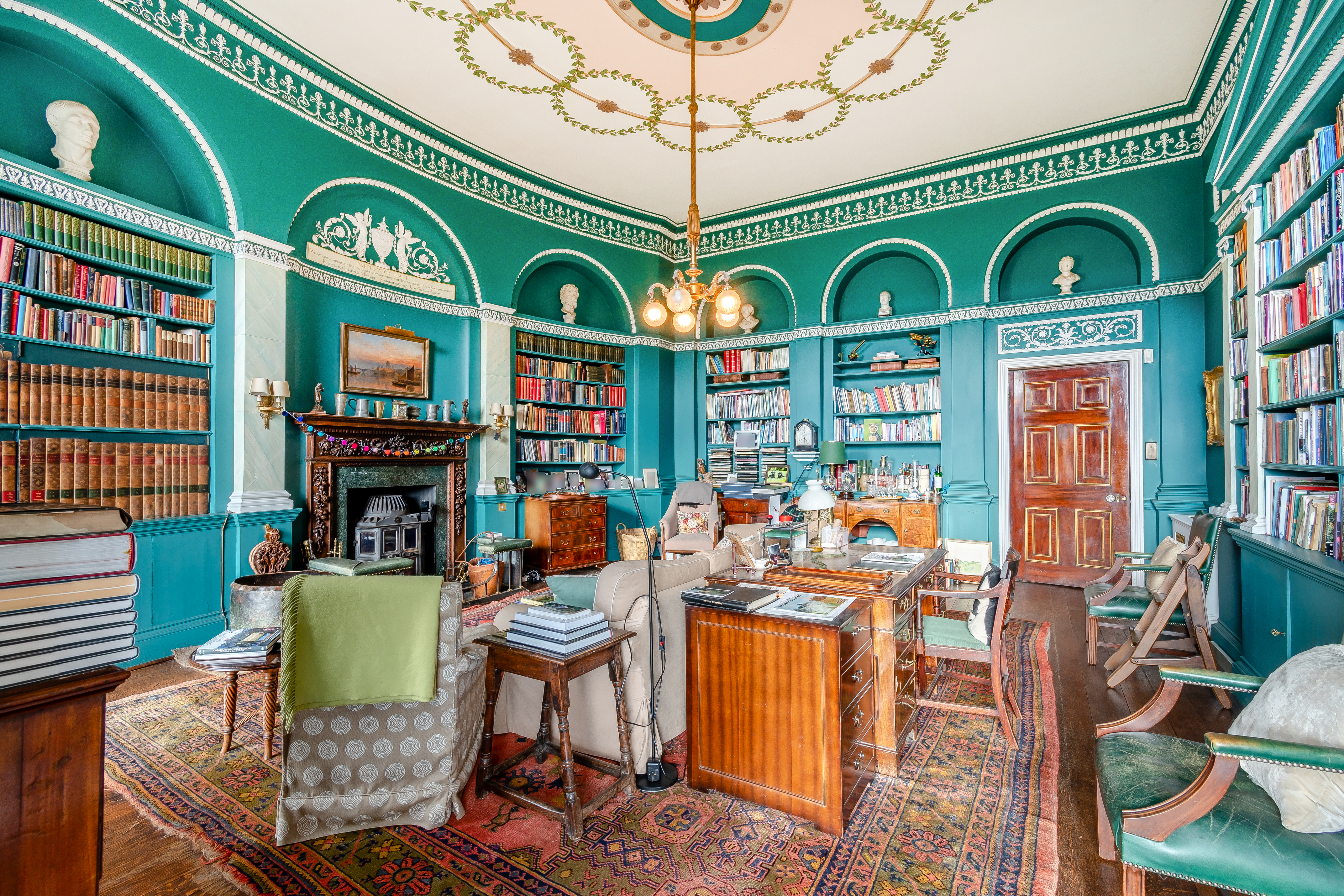 A teal library with an ornate ceiling and many colourful books and pieces of furniture scattered about