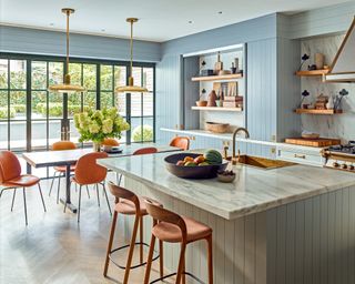 kitchen dining area with island with marble top, dining table with orange dining chairs and gold pendant lights