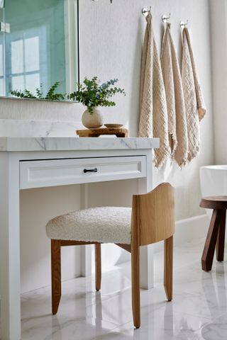 White bathroom with narrow white vanity with marble countertop and wooden chair with boucle fabric