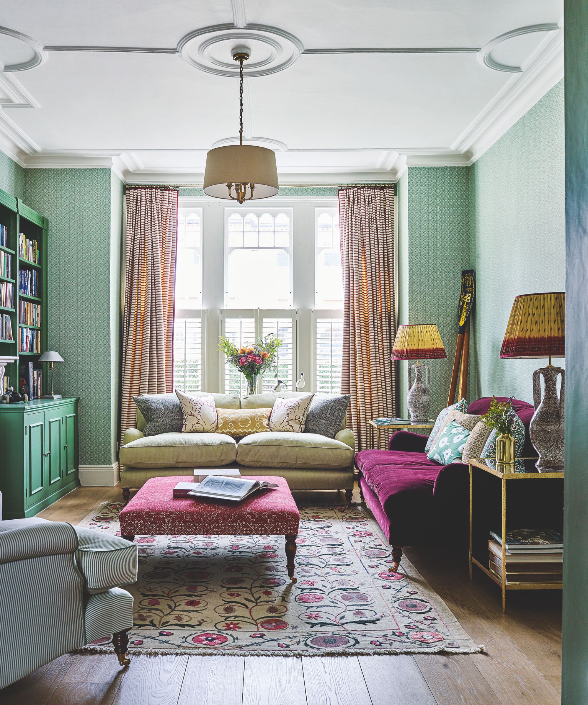 A cozy green living room with colorful layered patterns. In the centre of the room is a pink patterned ottoman, with a green couch and a dark pink velvet couch, on top of a large floral rug. To the back of the room are large windows with white shutters and striped curtains, and a large vase of flowers. To the left are green cabinets and shelves, and in the foreground is a green and white striped upholstered chair.