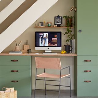green cupboards and drawers with chrome handles with a desk with a Apple computer and office products, and pink chair in under stairs office set up
