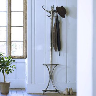 Henry Wall Mounted Coat Stand finished in tarnished silver, with a coat, hat and umbrella hanging up, and located in a room with white walls next to a window and plant