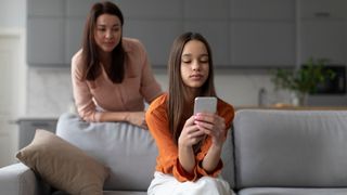 teenager looking at phone on a couch while her mother stands behind her