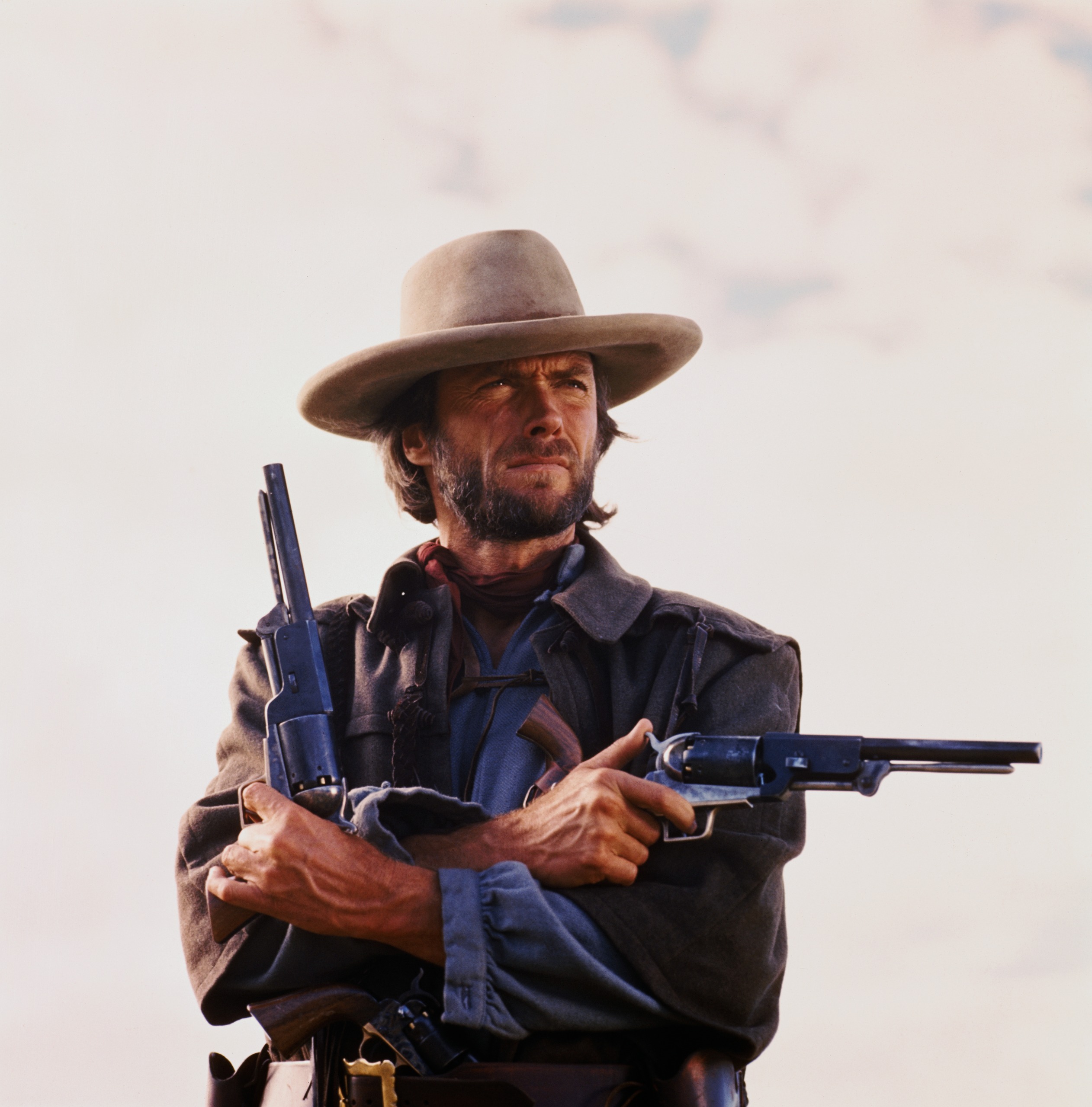 Clint Eastwood starring in the 1976 Western The Outlaw Josey Wales. Photo shows him from the waist-up holding two pistols, one pointing to his left and one pointing up, arms crossed.