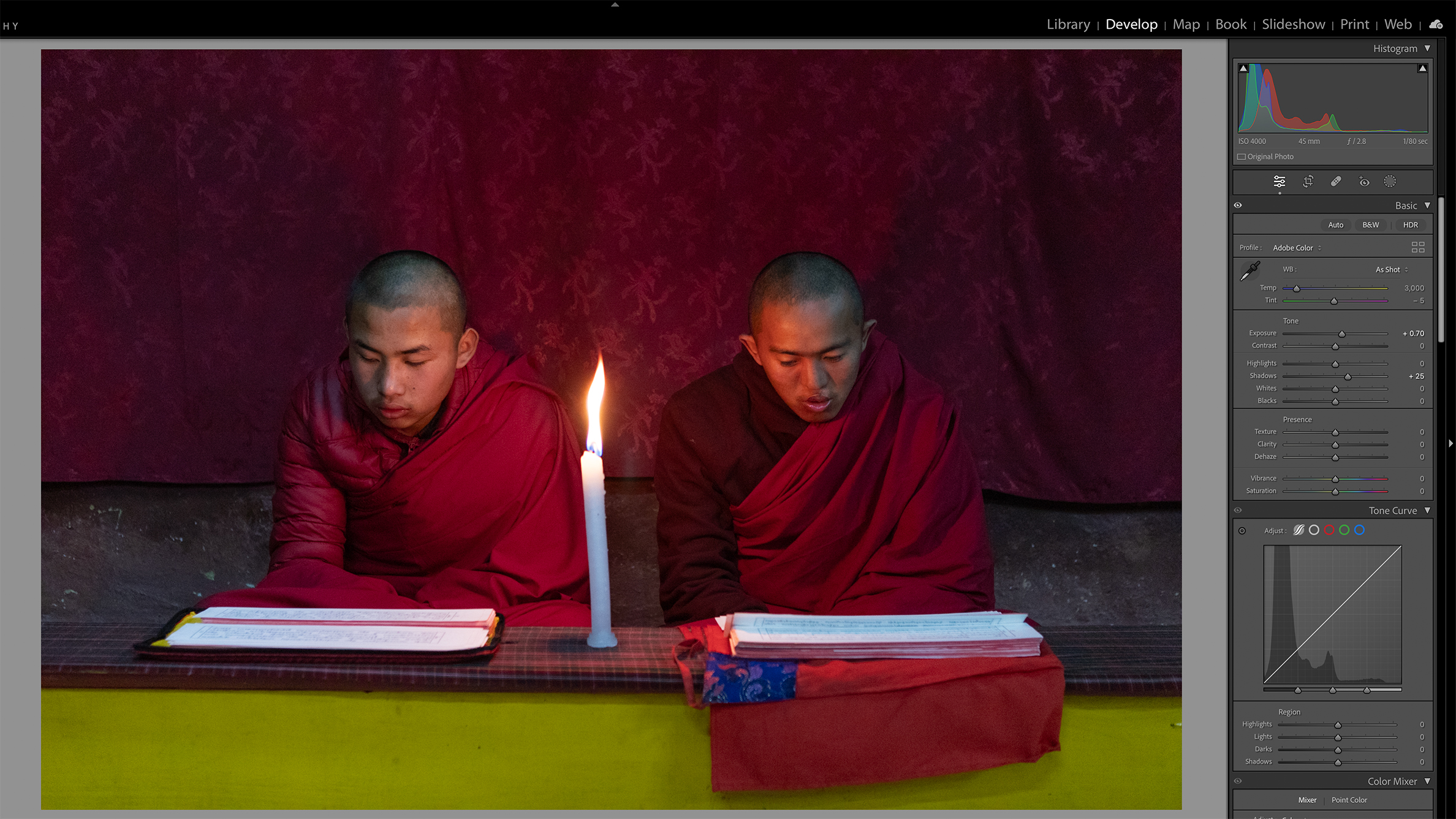 Two individuals in maroon robes are seated side by side, reading by candlelight. The dim setting conveys a calm and meditative atmosphere