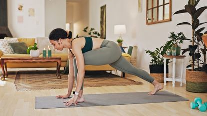 A woman practices a runner's lunge pose in a living room on a yoga mat. Her left knee is bent at a right angle, her hands planted either side, and her right leg is extended behind. In the background we see a couch, coffee table and candles. To her side are leafy plants and a pair of dumbbells.