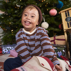 Child sat under a Christmas tree wearing a brown stripey top and brown beanie sold at Not on the high street