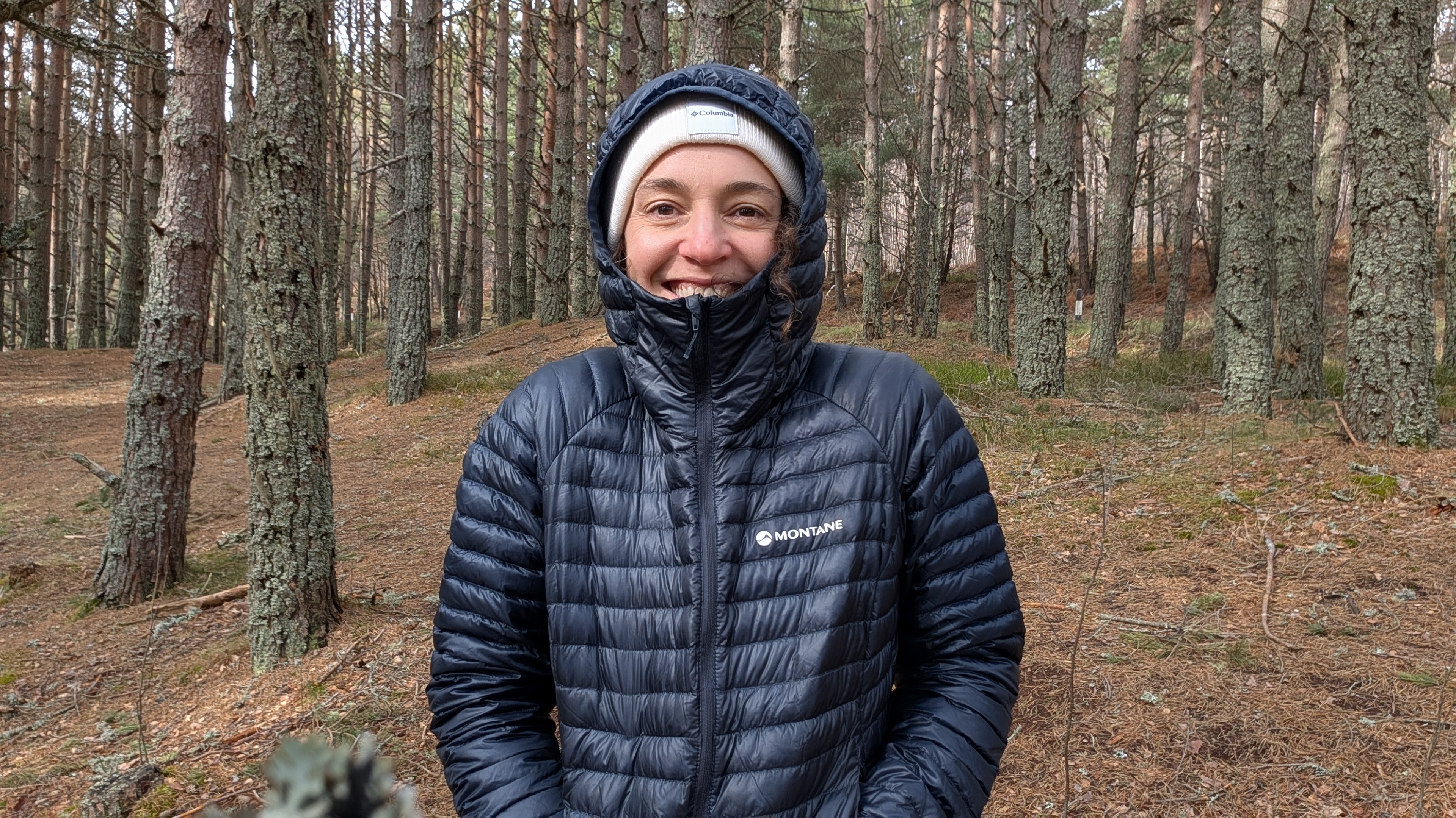 A woman in a forest wearing a dark blue down jacket with the hood up laughing and looking at the camera