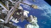 two astronauts in white spacesuits work near a large antenna dish outside a space station with a blue and white planet as their backdrop