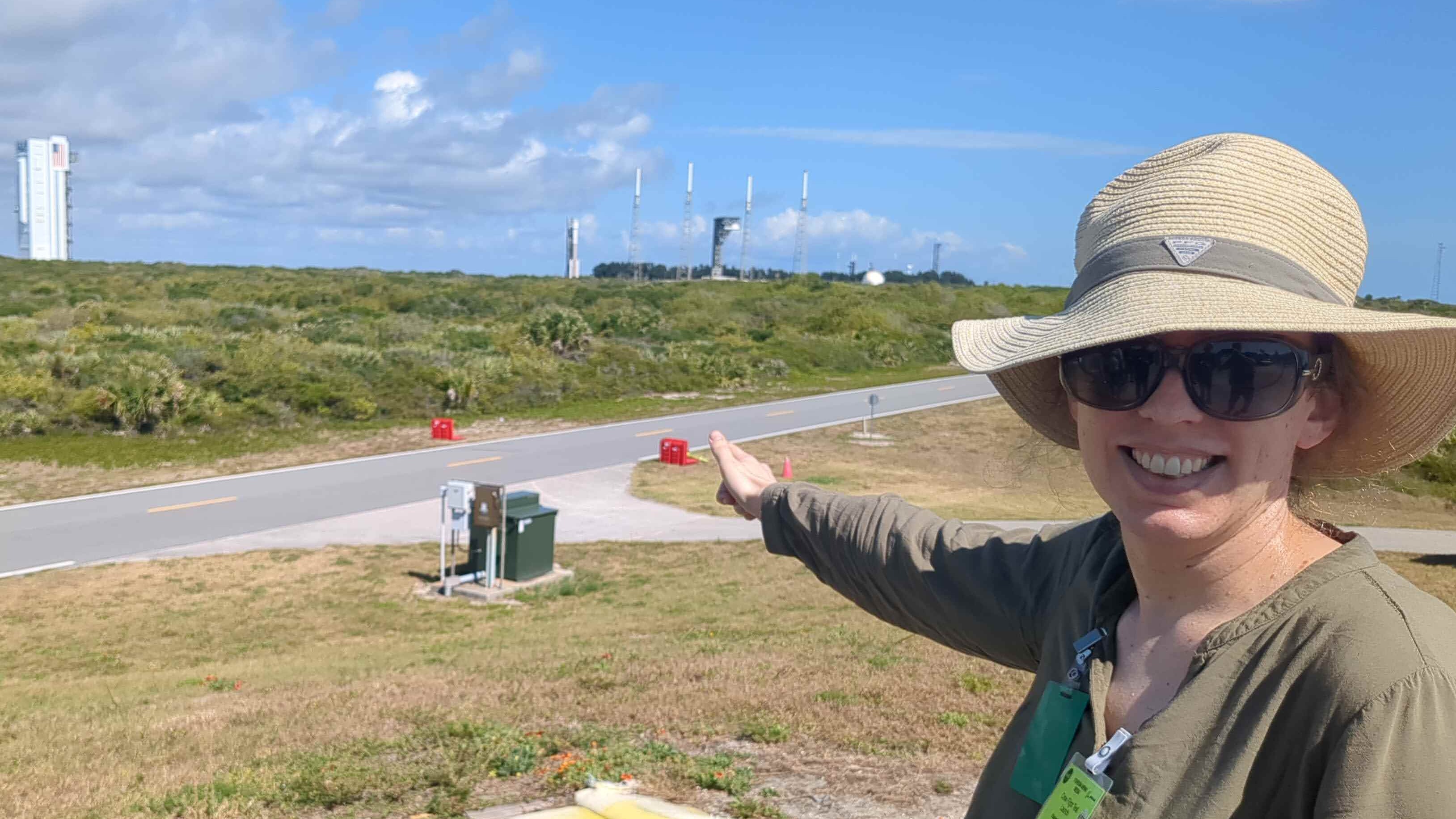 Wanita tersenyum mengenakan topi besar dan kacamata hitam.  Dia menunjuk ke seberang jalan pada sebuah roket dan landasan peluncuran di kejauhan sebagai latar belakang