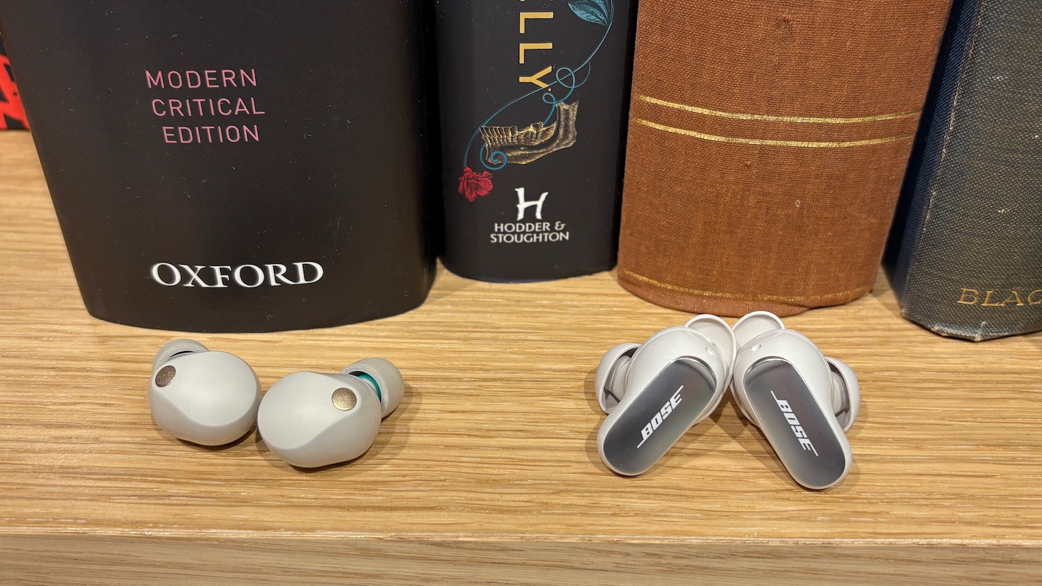 The Bose QuietComfort Ultra Earbuds (2nd Gen) and Sony WF-1000XM5 on a shelf in front of a row of books.