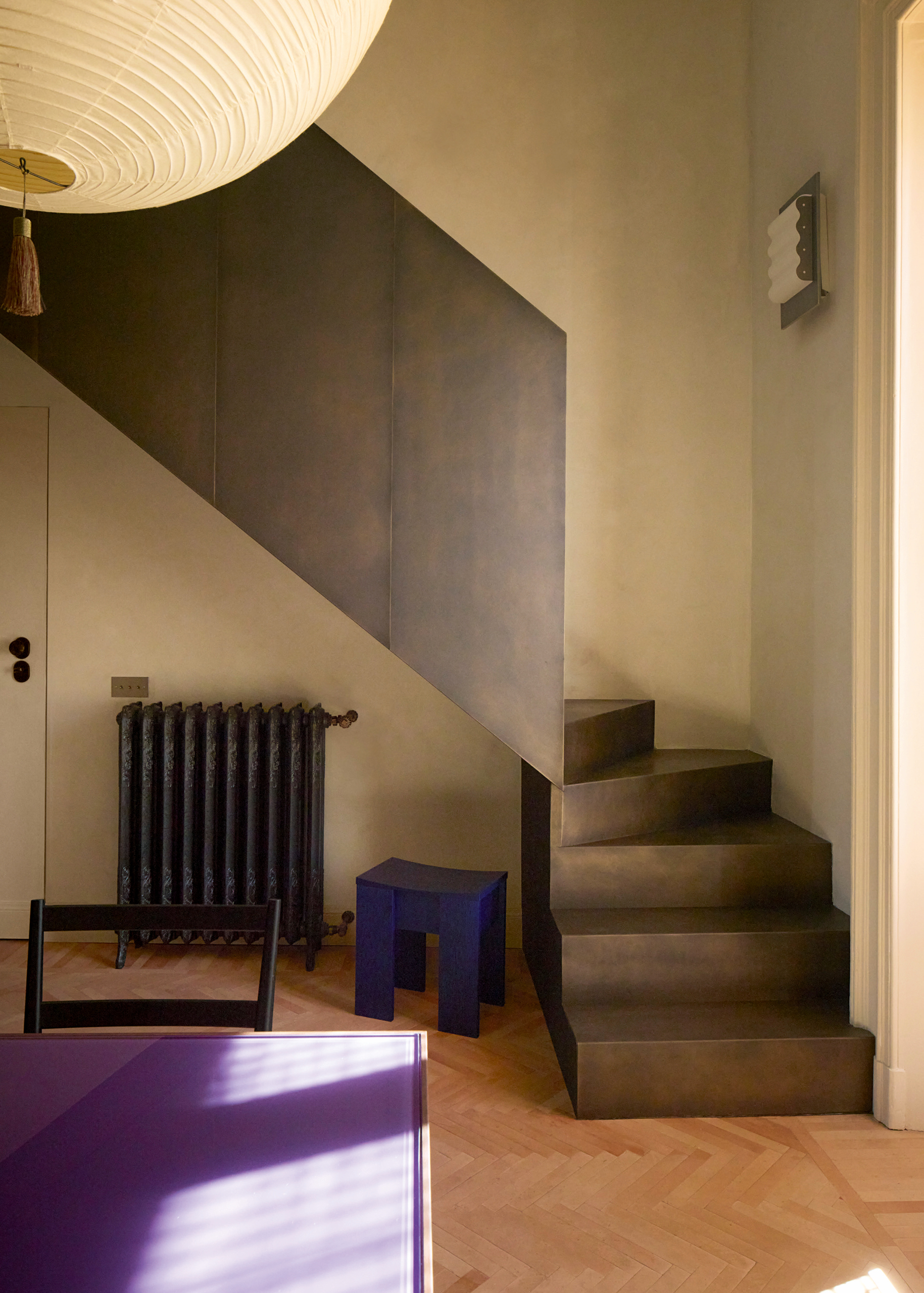 Hallway with purple table and black chair just in shot, blue stool, black case iron radiator, paper pendant, and a bronze angular enclosed staircase.