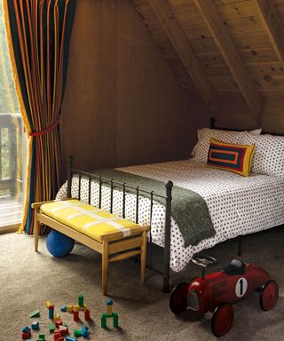 Kids bedroom with wooden paneling on walls, bed with patterned bedding, toy car, yellow upholstered bench at foot of bed