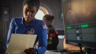 A woman with short black hair wearing a blue jumpsuit with an American flag patch on the shoulder is reading an important looking document. She looks concerned. In the background you see technical screens and some more space staff.