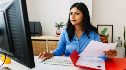 A woman looks at her computer screen and paperwork while sitting at her desk in an office.