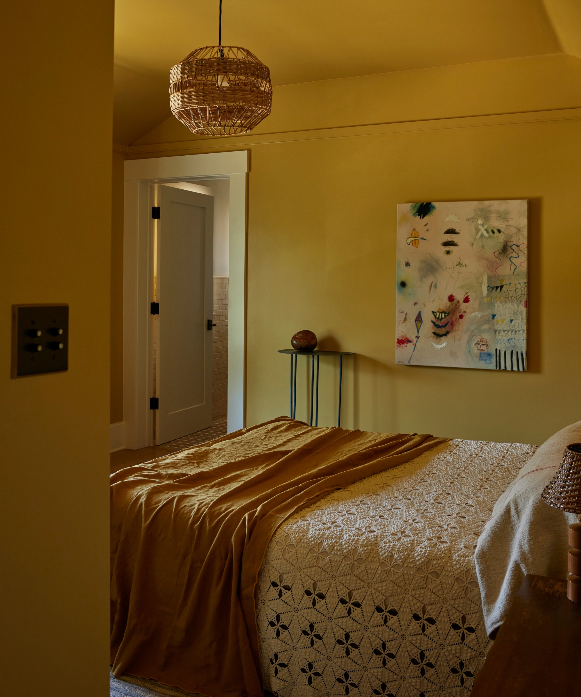 Ochre bedroom with matching walls and ceiling, patterned bedding, an amber throw, pendant light and colorful artwork.