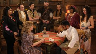 From left to right: Sam and Jay sitting at a table facing each other. Behind them, from left to right, Bela, Isaac, Pete, Thor, Sass, Alberta dn Flower all stand around the table.