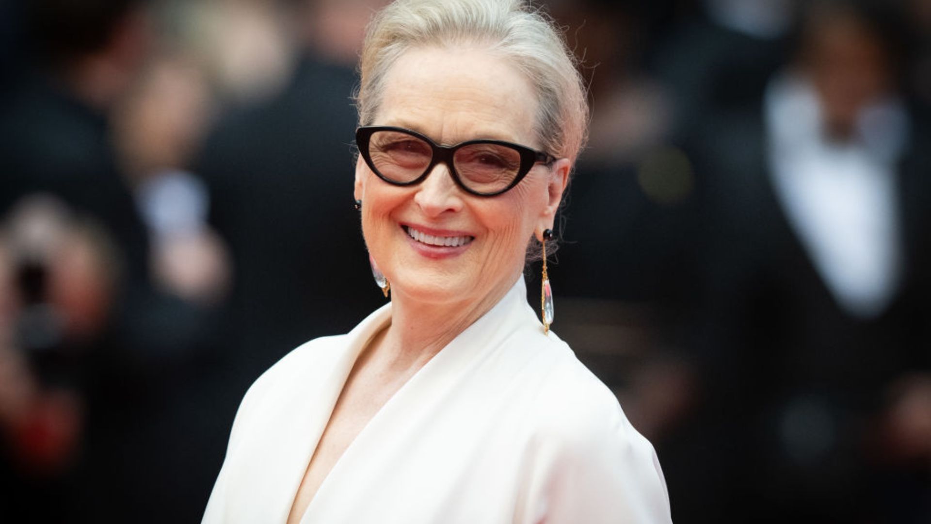 Meryl Streep smiles in a plunging cream blouse, drop earrings and black rimmed glasses