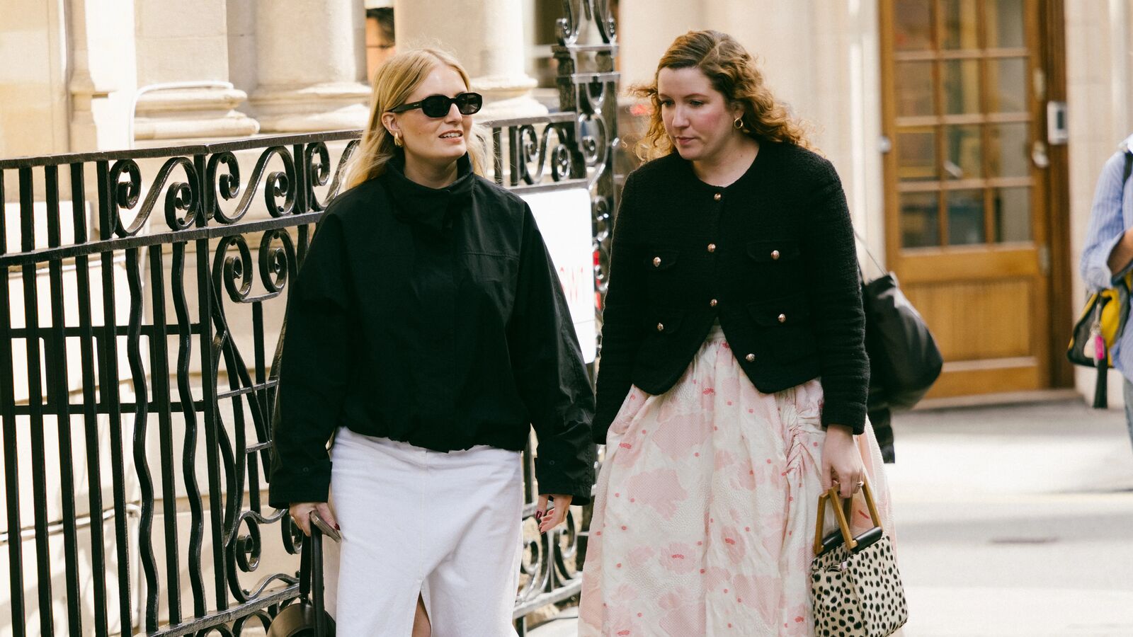 women walk the streets of london in fashionable outfits. one wears black sweater and white skirt while the other wears black cardigan sweater and fun pink skirt