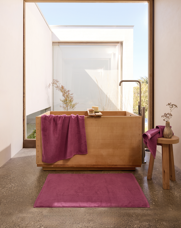 Image of a timber bathtub in a sunlit bathroom with magenta towels and a magenta bath mat.