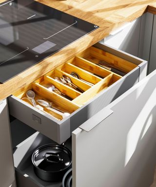 A modern kitchen drawer system with a wooden cutlery organiser is open beneath a black induction cooktop. The lower drawer holds black cookware, while sunlight casts soft shadows on the light grey cabinetry.