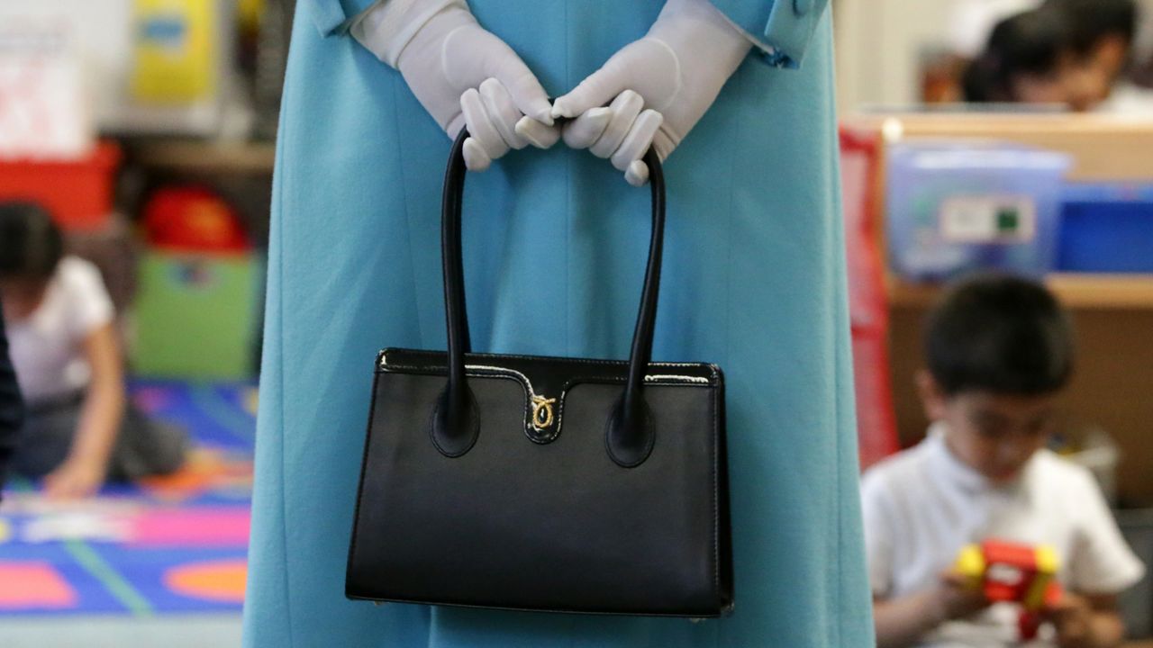 Queen Elizabeth II holding a black handbag behind her back and wearing white gloves with children playing in front of her