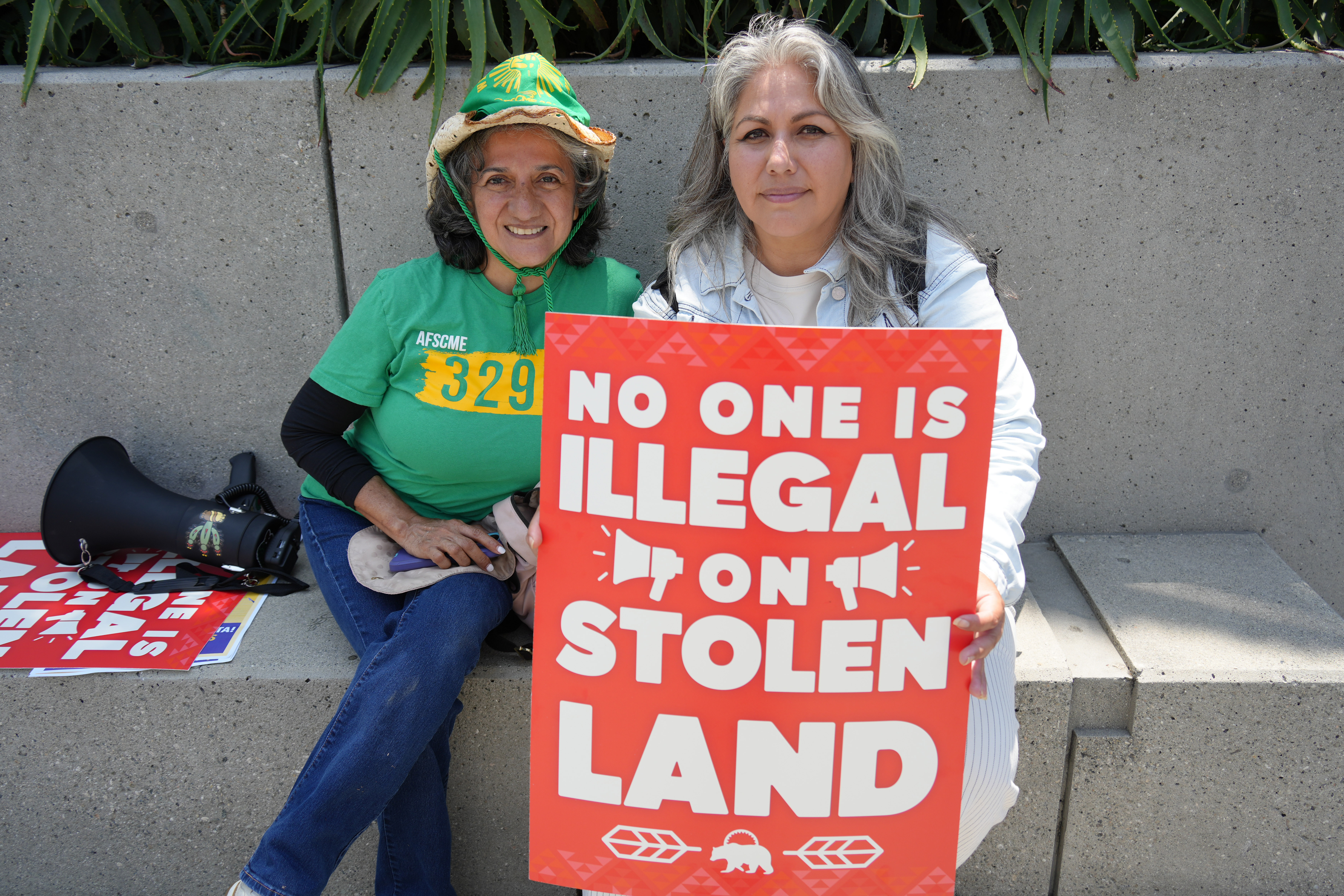 two women hold a red sign that reads “NO ONE IS ILLEGAL ON STOLEN LAND.”