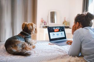 A woman using her laptop to browse Black Friday deals while laying on her bed with her dog.