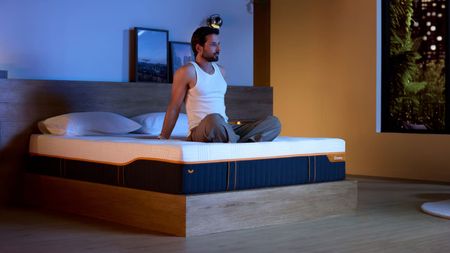 A man sits on an Emma Performance mattress on a bed frame in a dimly lit room