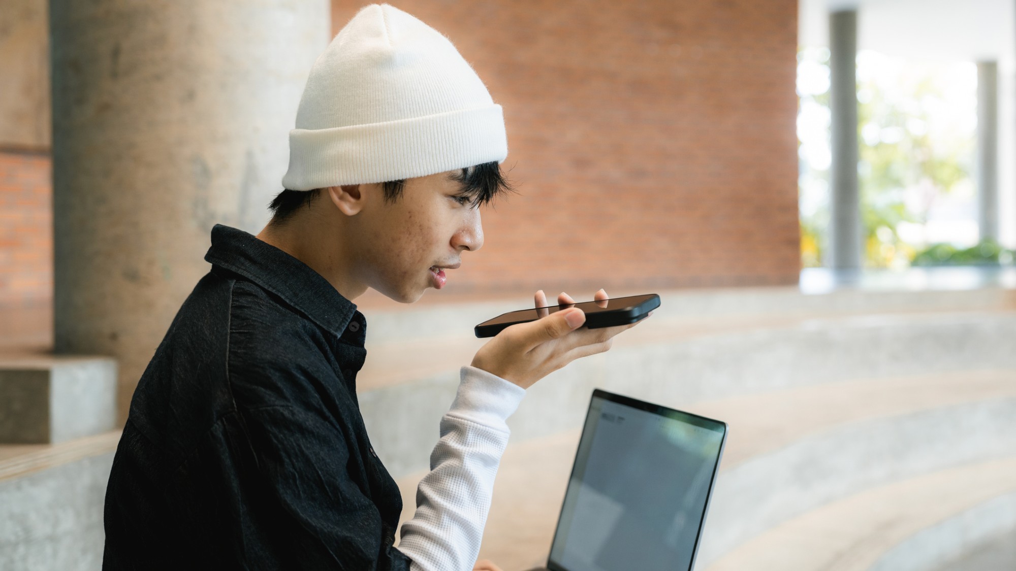 A young man speaks into a cell phone as he works on a laptop