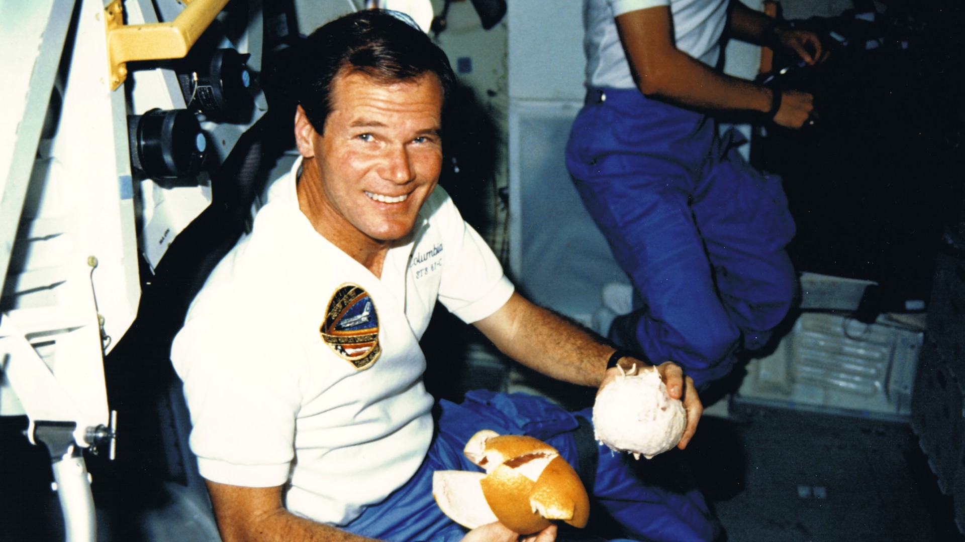 a man in a white three-button shirt and blue pants smiles while holding a peeled grapefruit in a cramped space shuttle cabin